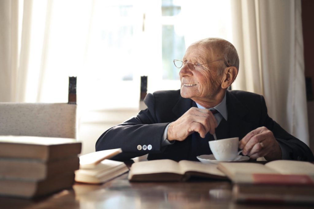 senior man drinking hot beverage while sitting at table with books