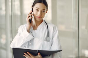 positive young doctor in medical uniform talking on cellphone in clinic corridor