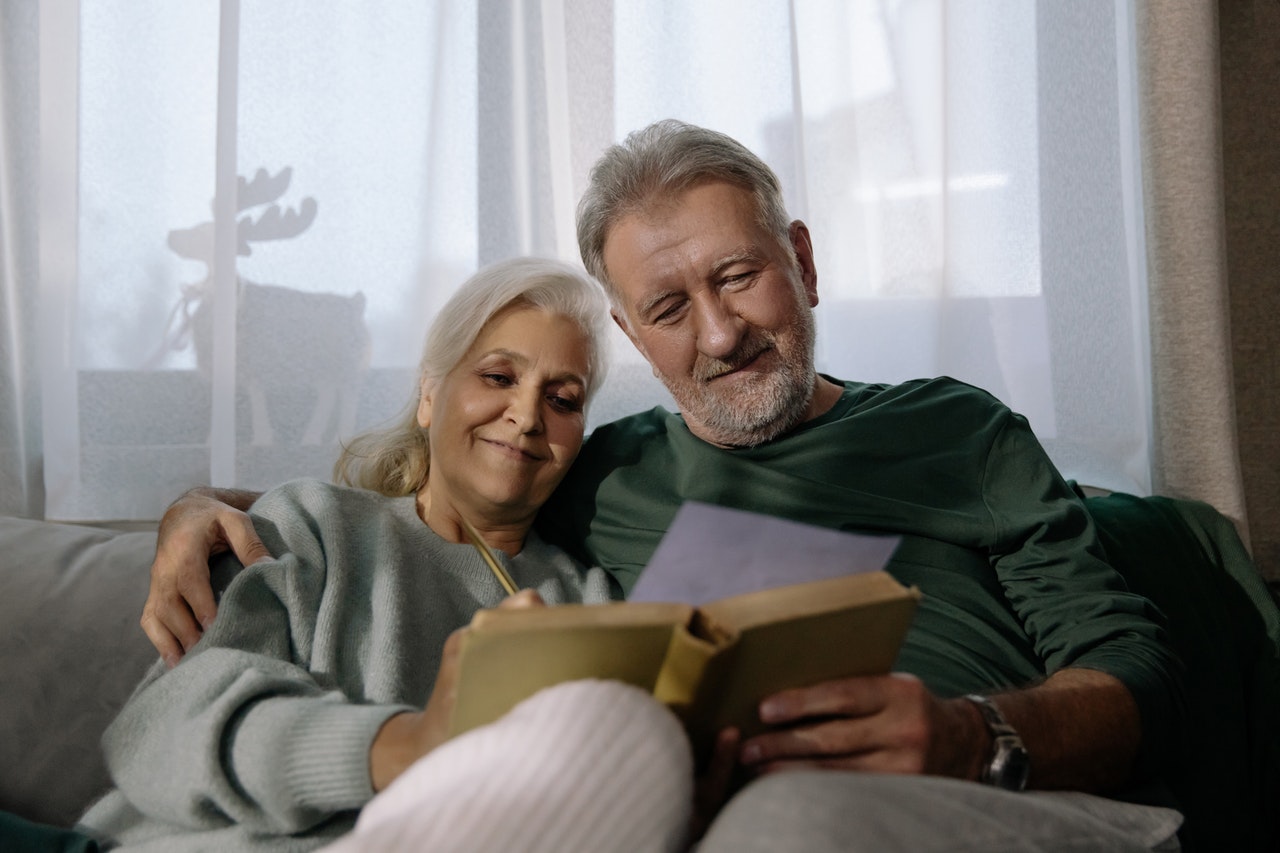 smiling older couple sitting together on the sofa with a book and notepaper