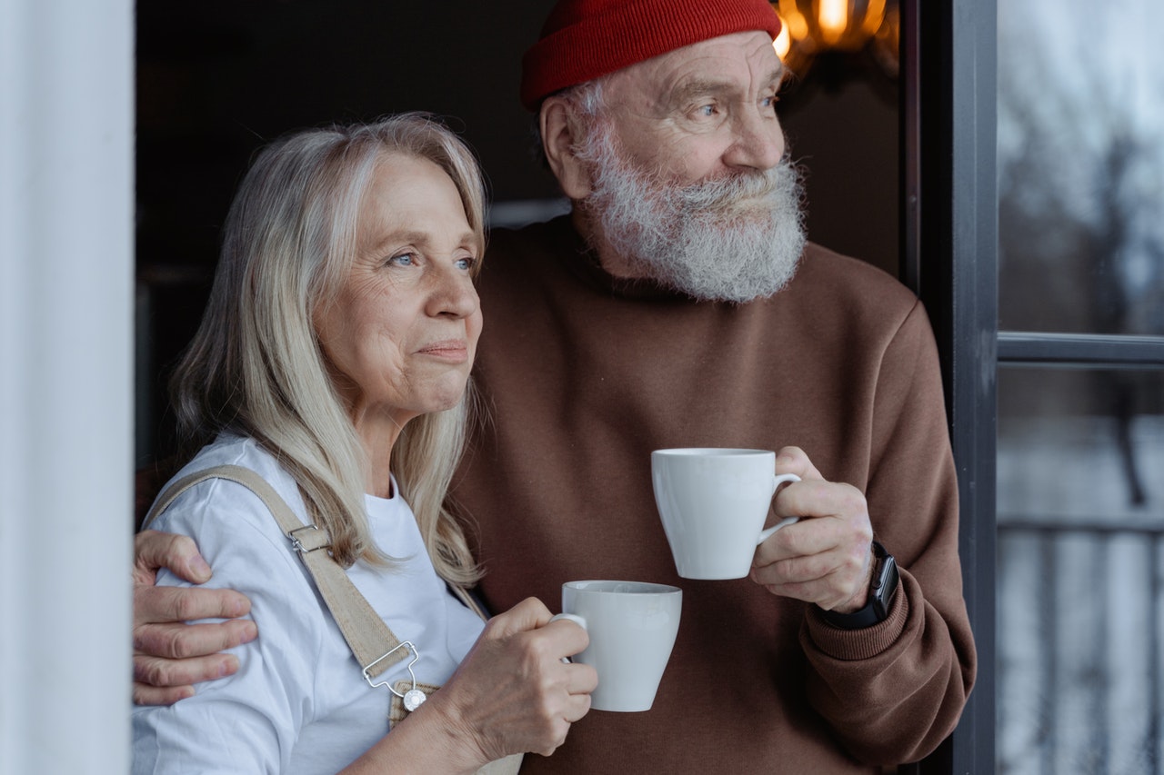 old couple holding mugs and looking out window together