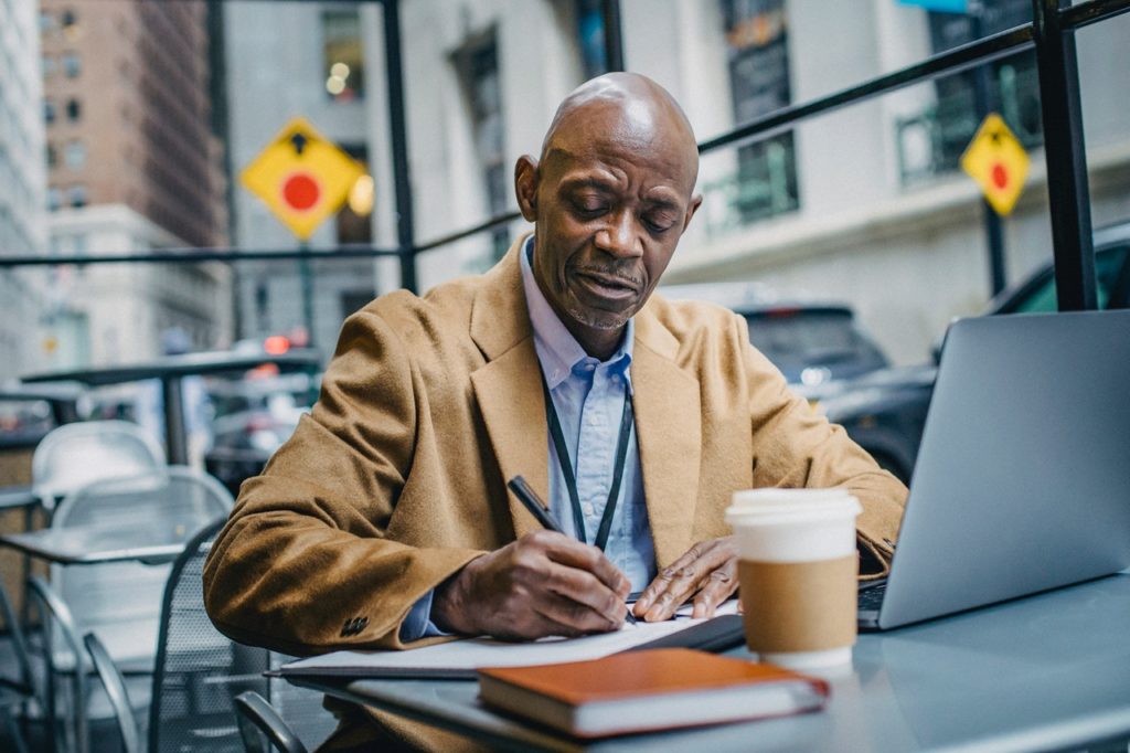 black man writing notes while working on laptop in coffee shop