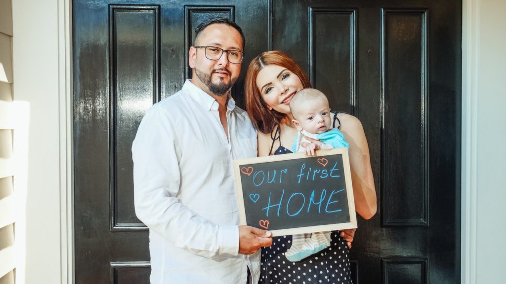 young couple holding baby with first home sign