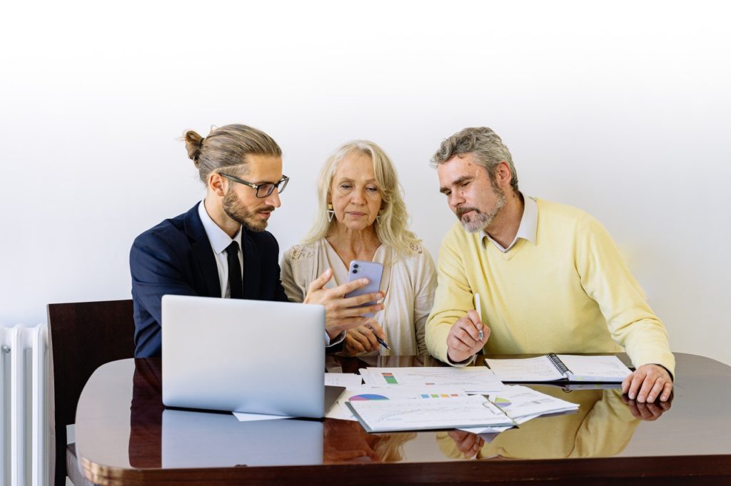 young businessman shows phone to older couple seated at conference table with laptop and paperwork