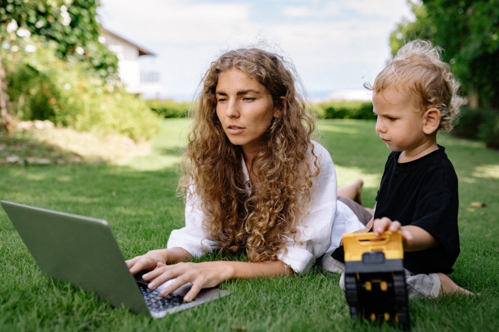 woman lying on grass with laptop and toddler with toy
