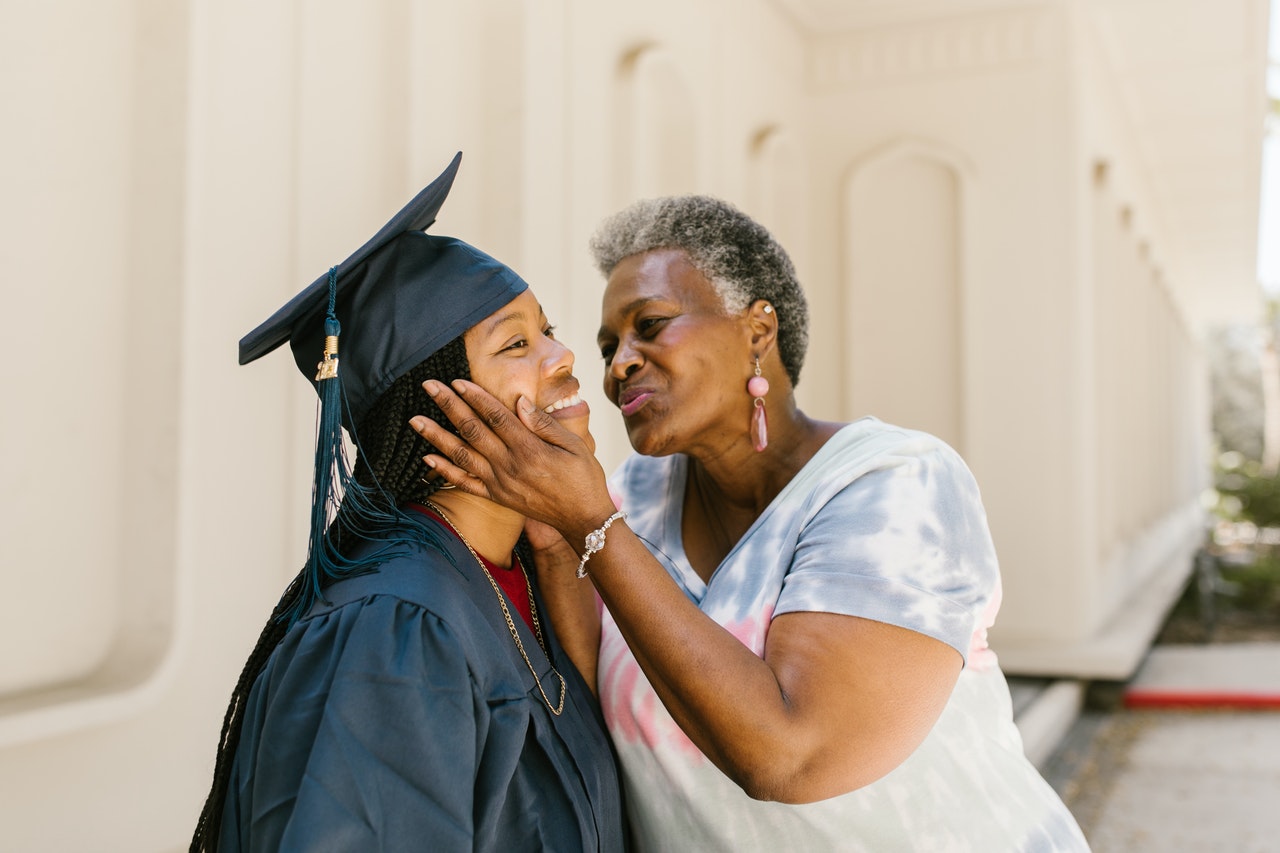 older black woman holding young female graduate's face for a congratulatory kiss on the cheek