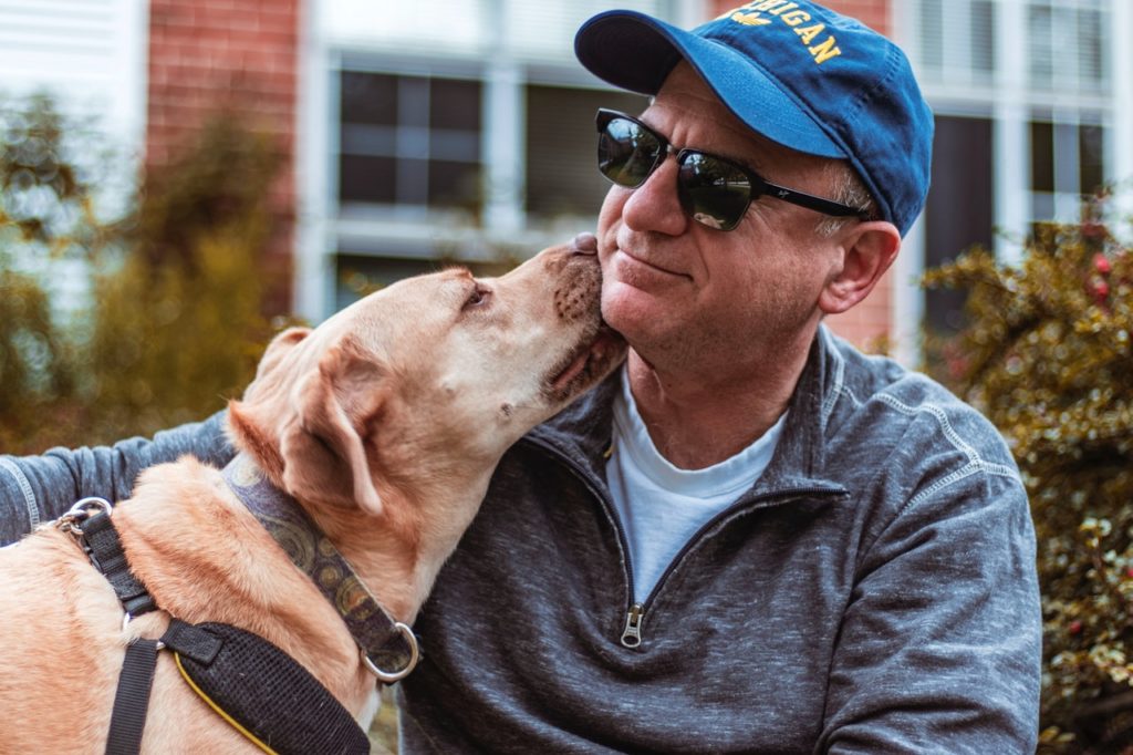 man in blue baseball hat sits outside with arm around dog as it licks his neck
