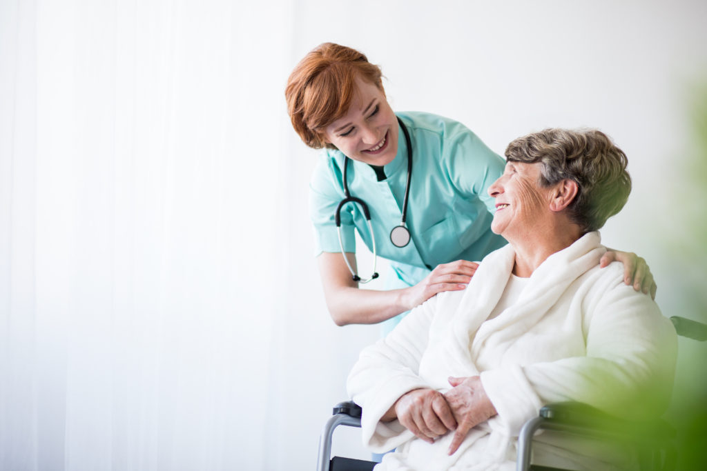 nurse smiles down at smiling elderly woman in a wheelchair