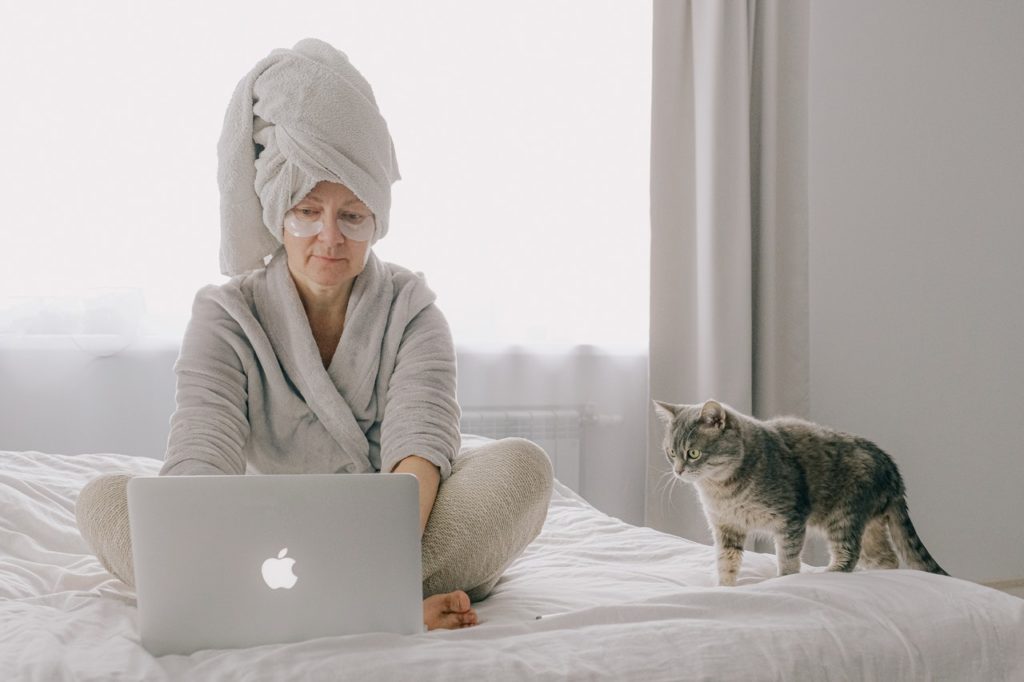 woman sits on bed in robe with towel on her head as she types on laptop with her cat nearby