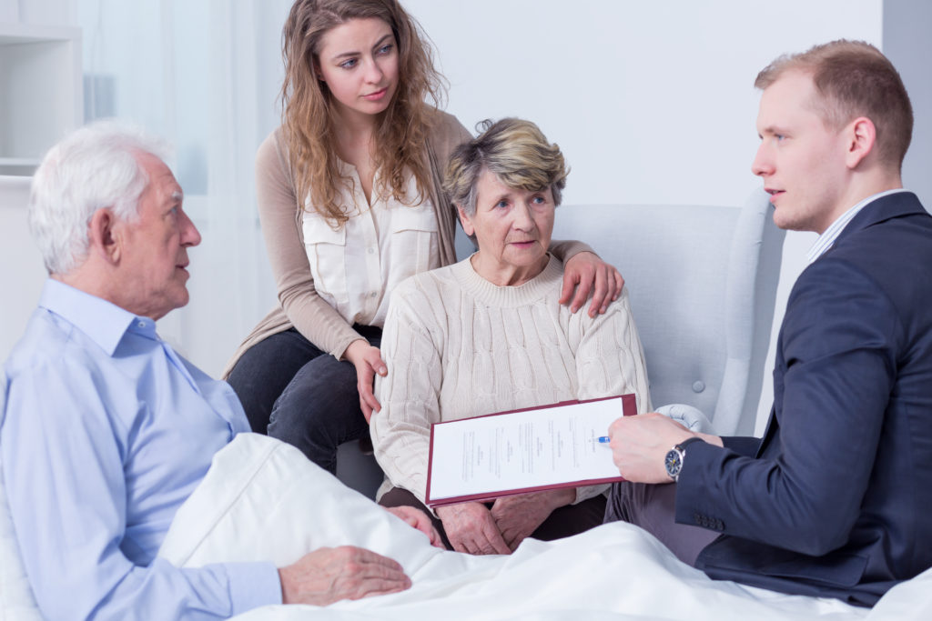 family sits beside an old man's hospital bed as they talk with a suited man holding a clipboard