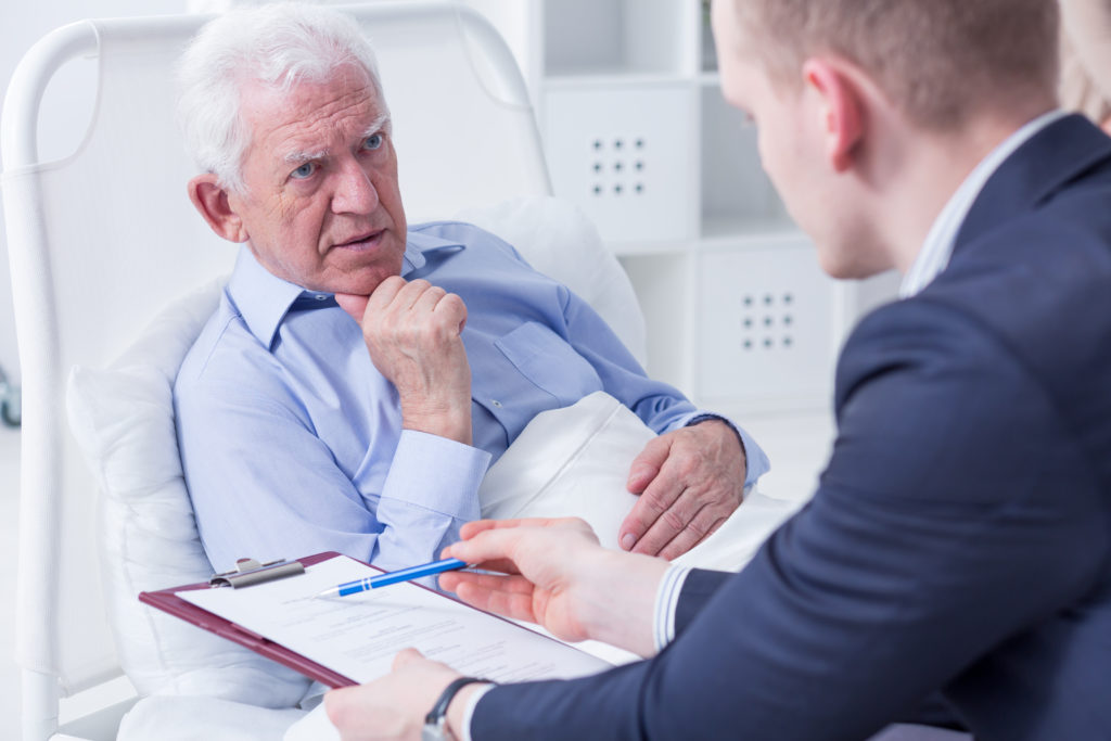 older man lies in a hospital bed while talking seriously with a man in a suit holding a clipboard